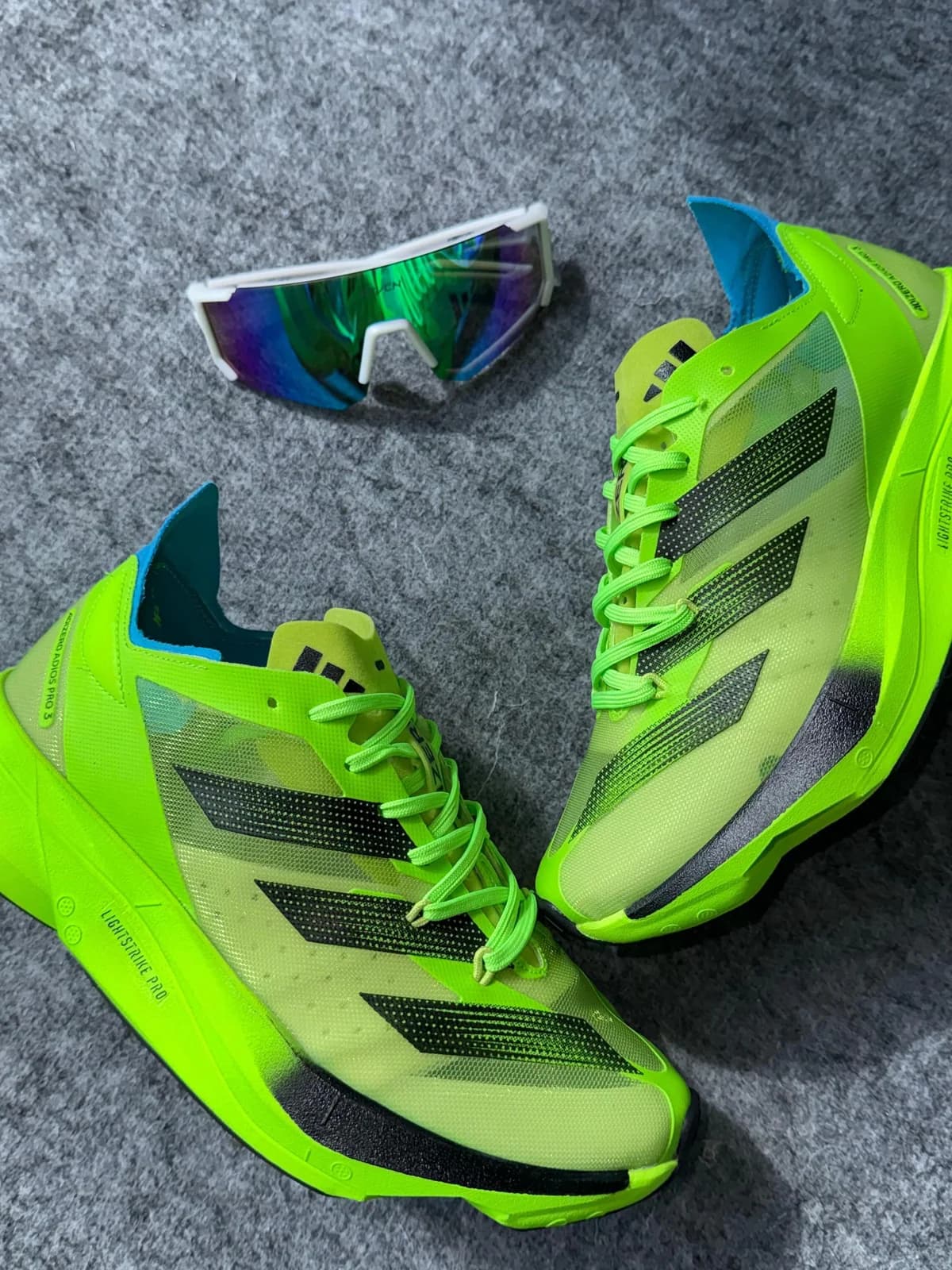 Dois pares de Adidas Adizero verde neon no carpete cinza, com um óculos esportivo espelhado ao lado